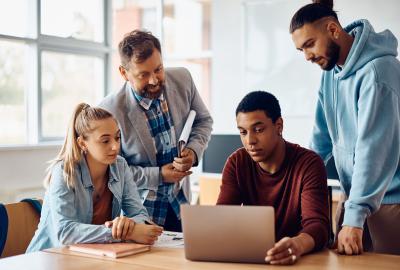 Group of learners gathered around a laptop with older man teaching them